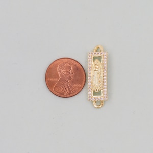 May include: Gold-tone charm featuring a depiction of the Virgin Mary with a clear crystal halo, surrounded by smaller clear crystals. The charm is rectangular and has a loop on the top for attaching to a bracelet or necklace.