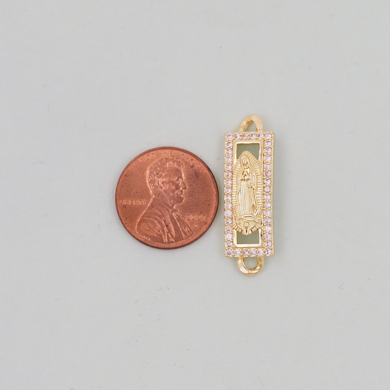 May include: Gold-tone charm featuring a depiction of the Virgin Mary with a clear crystal halo, surrounded by smaller clear crystals. The charm is rectangular and has a loop on the top for attaching to a bracelet or necklace.