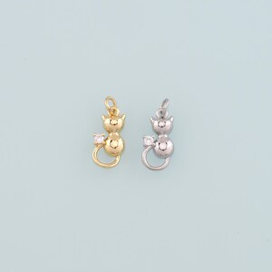 May include: Two cat-shaped charms, one gold-colored and one silver-colored, each with a small, clear gemstone. The charms have a loop at the top for hanging and a curled tail. The background is a light blue.