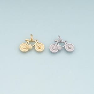 May include: Two small bicycle charms, one gold-toned and one silver-toned, are displayed against a light blue background. The charms are detailed with spokes and a classic bicycle frame design, perfect for jewelry making or crafting.