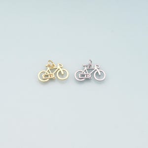 May include: Two bicycle charms, one gold-toned and one silver-toned, each with a small, clear accent. The bicycle charms are detailed with wheels, a frame, and handlebars. The charms are displayed on a light blue background.