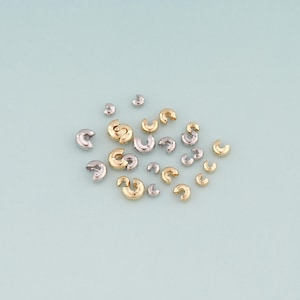 May include: A collection of gold and silver-colored crimp beads scattered on a light blue surface. The beads are crescent-shaped and vary in size, with a mix of shiny and matte finishes. These small jewelry findings are used for securing beads and other components.