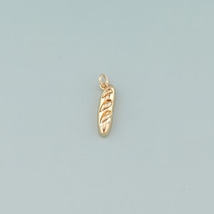 Könnte beinhalten: Ein goldfarbener Baguette-Anhänger mit strukturierter Oberfläche und einer kleinen Schlaufe zum Befestigen. Der Anhänger ist eine Miniatur-Darstellung eines Brotlaibs, ideal für Schmuck.