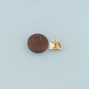 May include: A gold-colored envelope charm with a red heart detail. The charm is shown next to a penny for size comparison. The charm has a loop for attaching to a necklace or bracelet. The background is a light blue color.