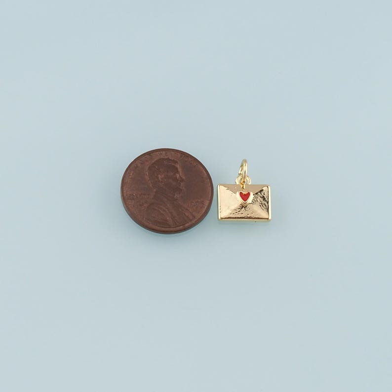 May include: A gold-colored envelope charm with a red heart detail. The charm is shown next to a penny for size comparison. The charm has a loop for attaching to a necklace or bracelet. The background is a light blue color.
