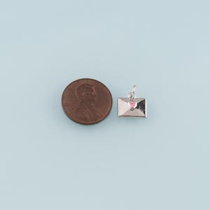 May include: A small silver envelope charm with a pink heart detail, next to a penny. The charm is likely for jewelry making or as a pendant. The background is a light blue.