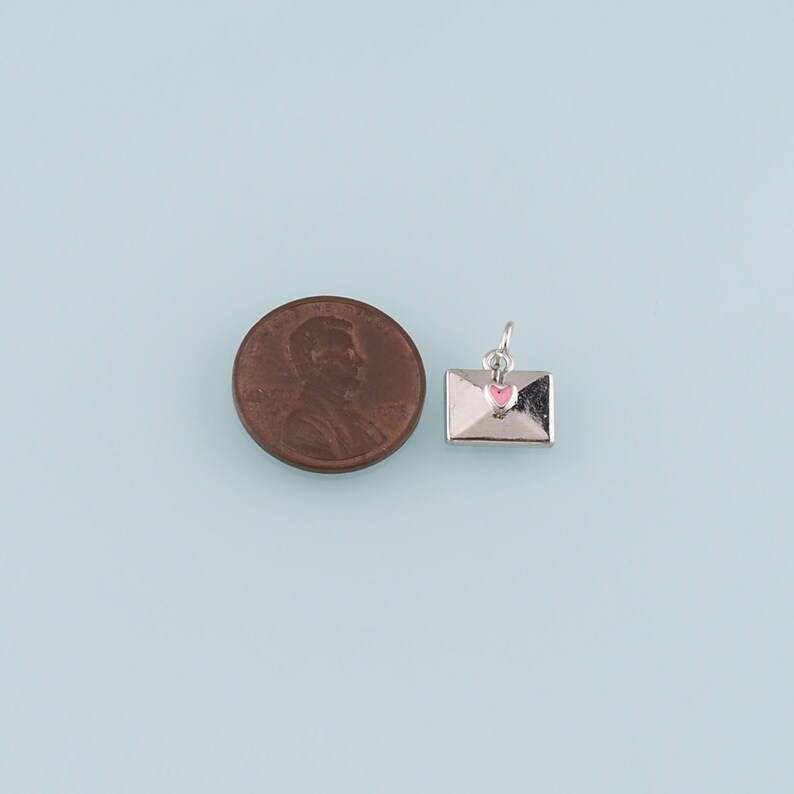 May include: A small silver envelope charm with a pink heart detail, next to a penny. The charm is likely for jewelry making or as a pendant. The background is a light blue.