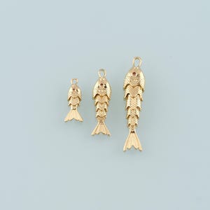 May include: Three gold-colored fish charms of varying sizes. Each charm features a detailed scale pattern, a small red gem for the eye, and a loop at the top for hanging. The fish tails are fanned out. The charms are displayed against a light blue background.