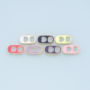 May include: Seven colourful enamel charms in various shades, including red, purple, and yellow, with a gold-coloured border. The charms are pull tab shaped, arranged on a light blue background.