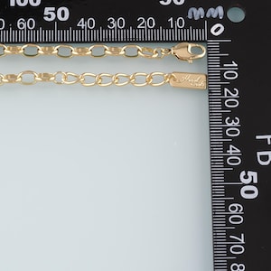 May include: A gold-colored chain necklace with an oval link design. The necklace has a lobster clasp closure and a small rectangular tag with the text "Hand Made". The necklace is measured against a ruler.
