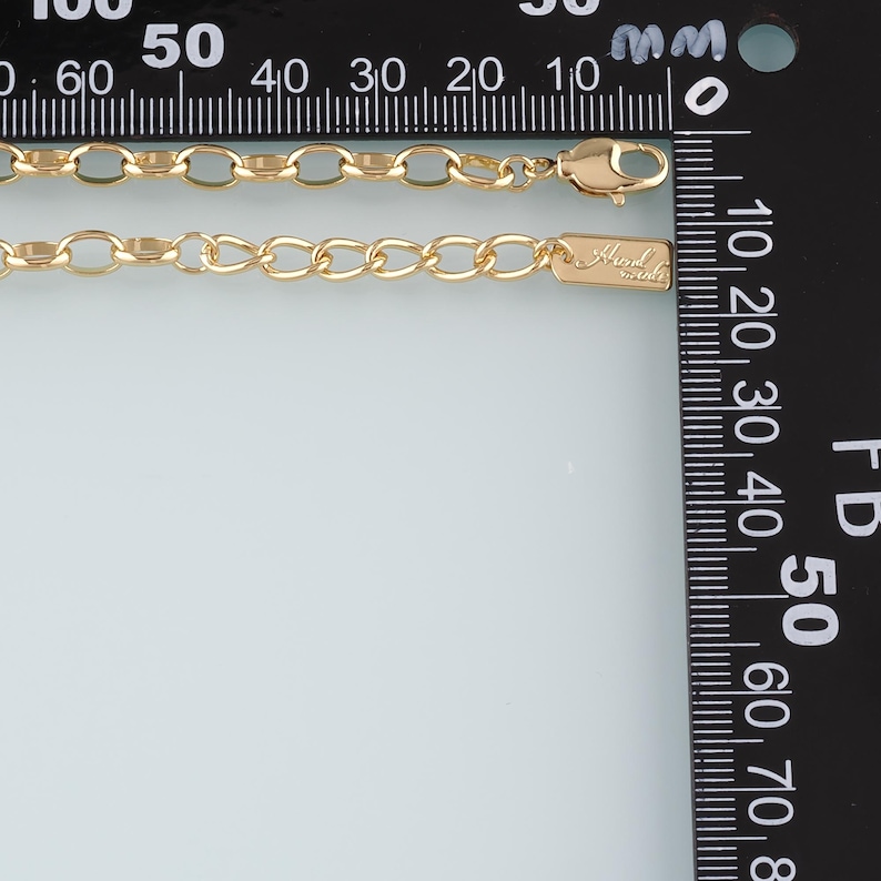 May include: A gold-colored chain necklace with an oval link design. The necklace has a lobster clasp closure and a small rectangular tag with the text "Hand Made". The necklace is measured against a ruler.