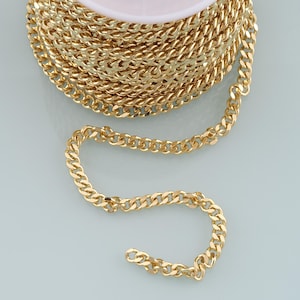 May include: A spool of gold-colored curb chain. The chain is made of small, interlocking links, creating a continuous, flexible strand. The chain is wound around a white spool, with a portion of the chain unwound and lying flat.