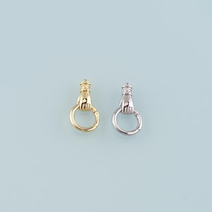 May include: Two metal clasp charms, one gold and one silver, each with a hand holding a ring, topped with a crown. The charms are set against a light blue background.