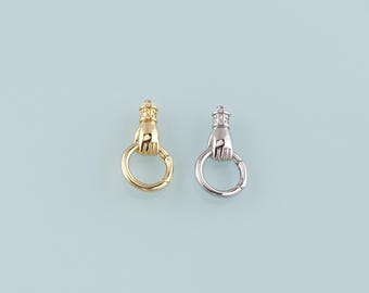 May include: Two metal clasp charms, one gold-toned and one silver-toned, each featuring a hand holding a ring. The hand is topped with a crown. The charms are set against a light blue background.