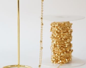 May include: A gold chain with small round beads, displayed on a gold stand. A spool of the same chain is visible in the background.