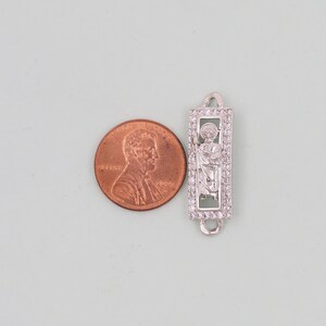 May include: Silver charm with a clear crystal border and a depiction of a saint inside. The charm is about the size of a UK penny.