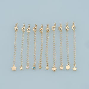 May include: A collection of gold-colored necklace extenders with various charms, including a flower, a small gem, a heart, and a star. The extenders are displayed against a light blue background.