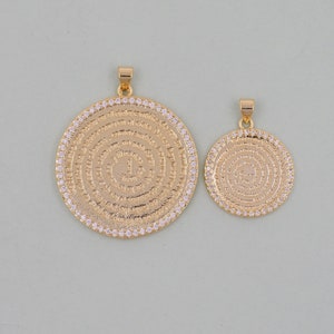 May include: Two gold-tone pendants with a circular design. Both pendants feature a circular pattern of small white stones and a central inscription. The larger pendant is slightly larger than the smaller pendant.