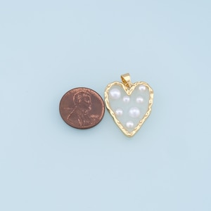 May include: Gold heart-shaped pendant with a white background and multiple white pearls inside. The pendant is about the size of a US penny.