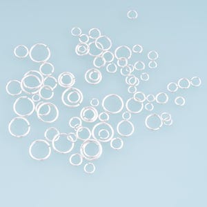 May include: A collection of numerous silver-colored metal jump rings scattered across the frame. Each ring features a circular shape with a small, visible opening or split, commonly used as jewelry findings for connecting various components.
