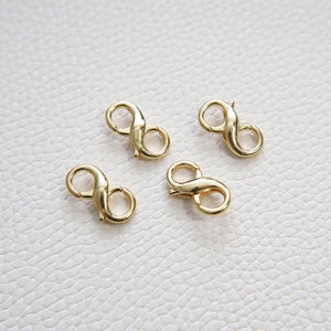 May include: Four gold-toned metal lobster clasps, each shaped like an infinity symbol, are arranged on a white textured surface.