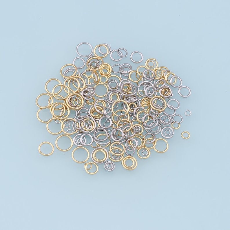 May include: A large pile of silver and gold jump rings. The rings are of various sizes and are scattered on a light blue background.