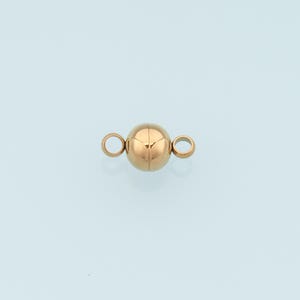 May include: A rose gold-toned spherical magnetic jewelry clasp featuring a smooth, polished surface. The clasp consists of a central ball-shaped magnet with two small, circular metal loops attached to opposite sides for connecting jewelry strands.