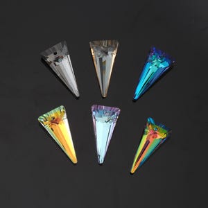 May include: Six iridescent, triangular crystal pendants in various colours, including silver, gold, blue, and rainbow hues. Each pendant is faceted, catching and reflecting light. The crystals are arranged on a dark surface.