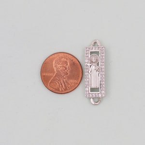 May include: Silver charm with a clear crystal border and a depiction of a saint holding a cross. The charm is about the size of a UK penny.