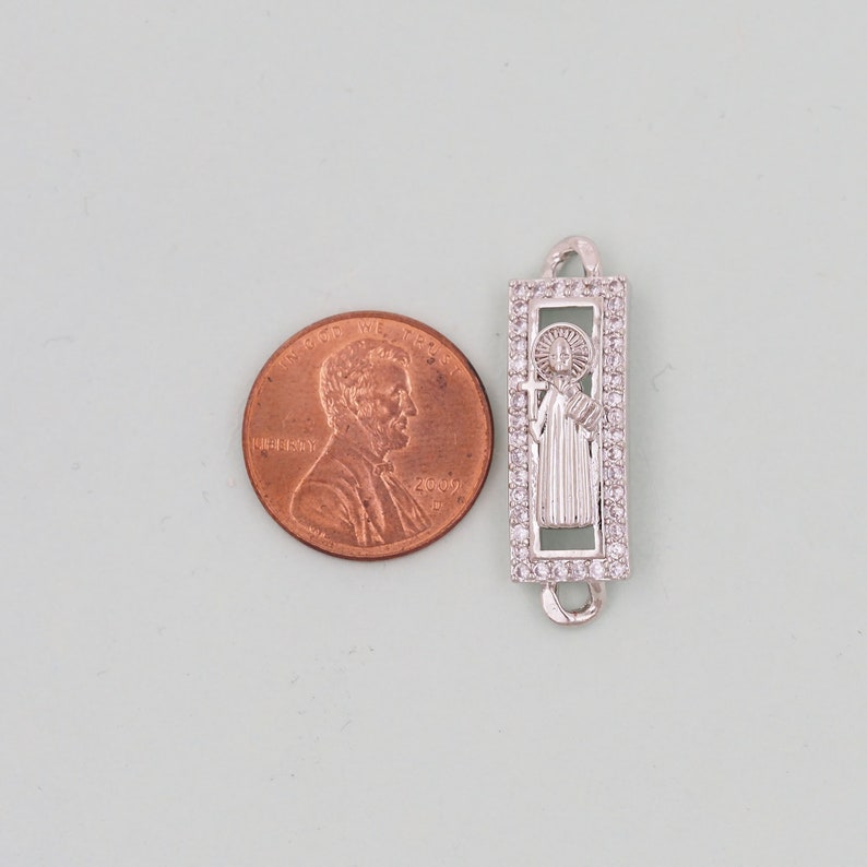 May include: Silver charm with a clear crystal border and a depiction of a saint holding a cross. The charm is about the size of a UK penny.