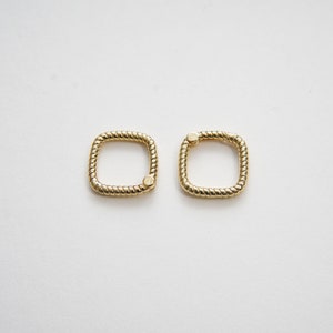 May include: Two gold-colored, square-shaped metal rings. Each ring has a twisted rope-like texture around the edges. A small, circular clasp is visible on one side of each ring. The background is plain white.