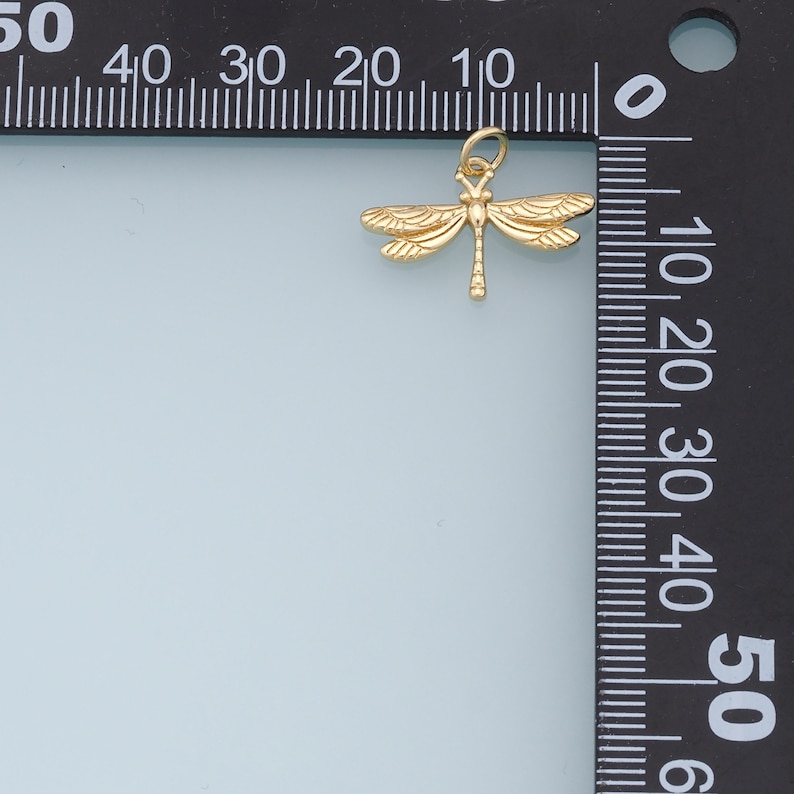 May include: A gold-colored dragonfly charm pendant, measuring approximately 1 cm in length, is shown against a white background. The charm is positioned next to a ruler, with the dragonfly's wings extending slightly past the 1 cm mark.