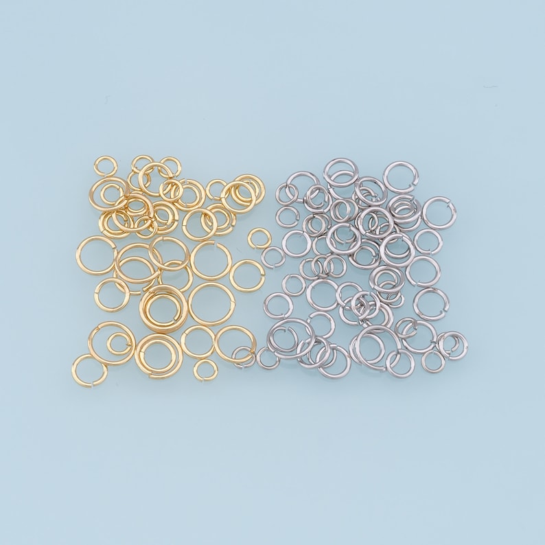 May include: A collection of gold and silver jump rings. The jump rings are arranged in a pile on a light blue background.