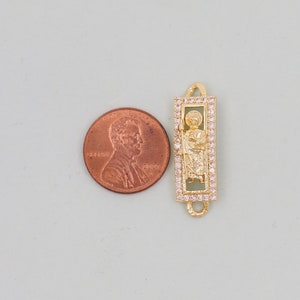 May include: Gold-tone charm with a clear crystal-encrusted frame featuring a religious figure. The charm is approximately the size of a UK penny.