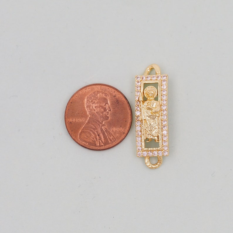 May include: Gold-tone charm with a clear crystal-encrusted frame featuring a religious figure. The charm is approximately the size of a UK penny.