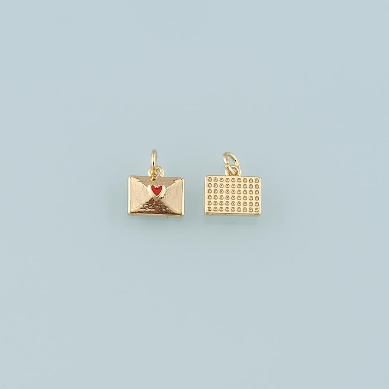May include: Two gold-colored envelope charms with a textured finish. One charm features a red heart, while the other has a grid pattern. Both charms have a small loop for attaching to jewelry. The background is a light blue.