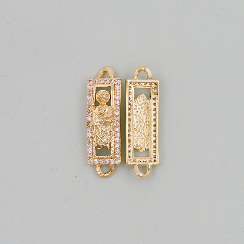 May include: Two gold-toned metal charms in a rectangular shape. One charm features a religious figure in a frame with clear stones. The other charm has a textured design.
