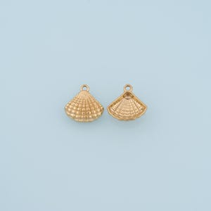 Op de afbeelding: Twee goudkleurige metalen bedels in de vorm van een schelp, elk voorzien van een klein rond oogje aan de bovenkant voor bevestiging. De linkerbedel toont de textuur van de geribbelde buitenkant van een schelp, de rechter de gladde binnenkant.