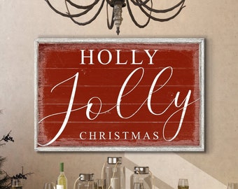 May include: Red wooden sign with white text that reads "HOLLY jolly CHRISTMAS". The sign is hanging on a wall above a wooden table set with white plates and wine glasses.