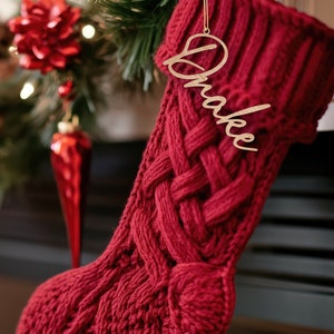 May include: Red knitted Christmas stocking with a wooden name tag that says "Drake" hanging on a fireplace mantel.