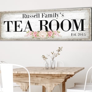 May include: A white wooden sign with black text that reads "Russell Family's Tea Room Est. 2015". The sign has a distressed look and is decorated with pink flowers.