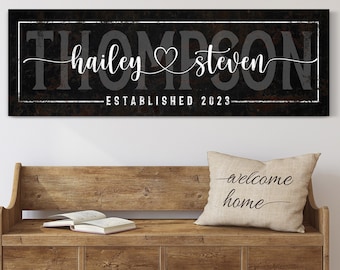 May include: A family name sign with the name "Thompson" in large, grey letters. The names "hailey" and "steven" are in white script, with a heart between them. The sign reads "ESTABLISHED 2023". A pillow with "welcome home" is on a wooden bench.
