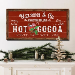 Op de afbeelding: Een rood houten bord met witte tekst die "Nelsons & Co. Christmas Blend Hot Cocoa Served Daily With Love" luidt. Het bord heeft een rustieke uitstraling en toont een cartoonafbeelding van een mok warme chocolademelk met marshmallows, snoepstokken en kaneelstokjes.