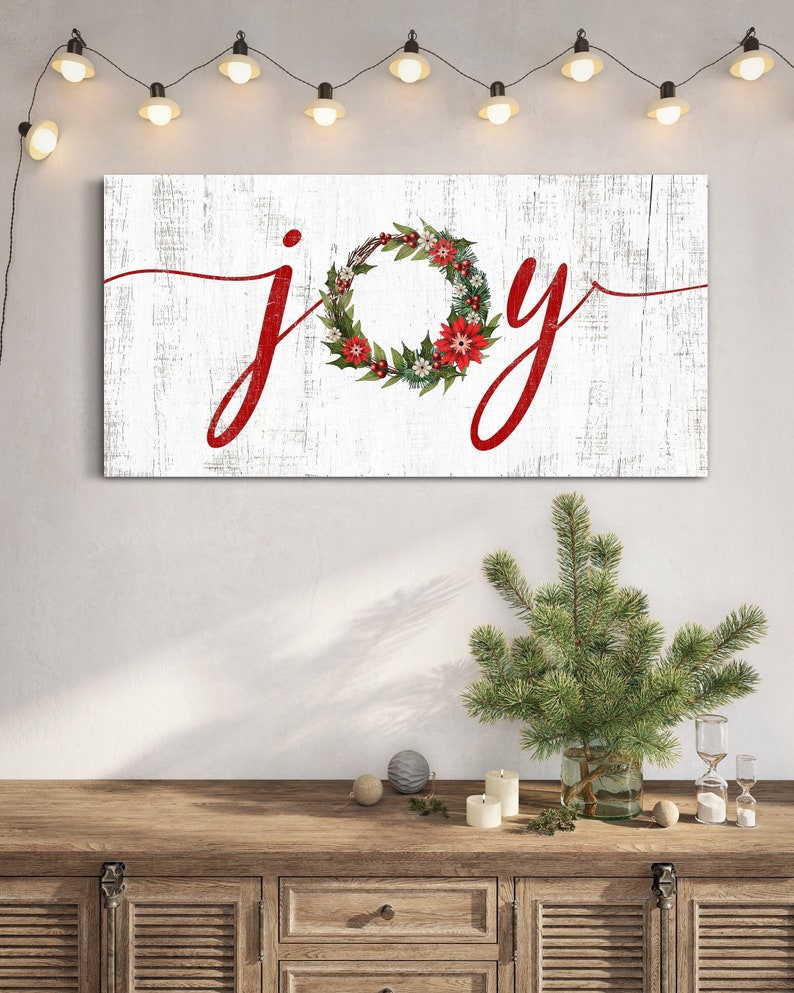 May include: White wooden sign with the word "joy" written in red cursive. The "o" in joy is a red and green Christmas wreath with red berries and poinsettia flowers.