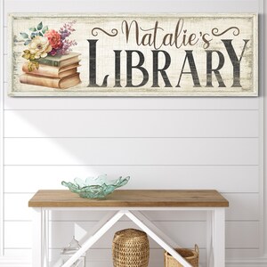 Pu&ograve; includere: Un cartello in legno bianco con una finitura usurata, decorato con un motivo floreale e il testo "Natalie's LIBRARY".