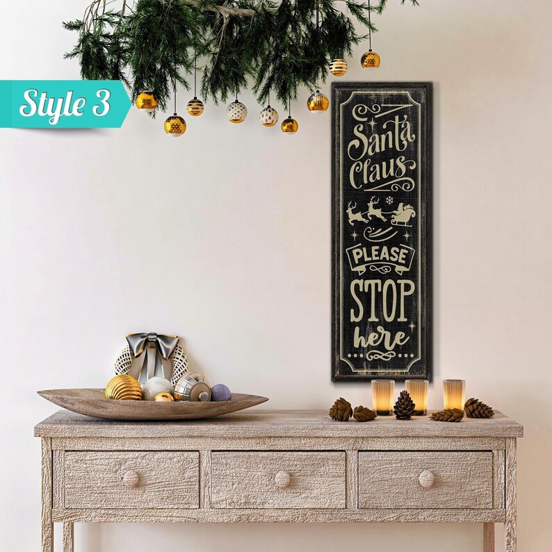 May include: Black and white sign with the text "Santa Claus Please Stop Here" in a vintage style font. The sign is hanging on a white wall with a wooden table and Christmas decorations in front of it.