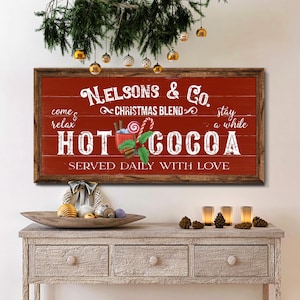 Op de afbeelding: Een rood houten bord met witte tekst die "come & relax" en "stay a while" luidt. Het bord toont ook de tekst "Nelsons & Co. Christmas Blend" en "HOT COCOA" met een afbeelding van een mok warme chocolademelk met slagroom, een snoepstok en kaneelstokjes. Het bord luidt "Served Daily With Love".