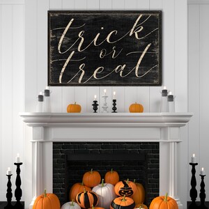 K&ouml;nnte beinhalten: Ein wei&szlig;er Kaminsims mit einem schwarzen Ziegelkamin und einem Schild mit der Aufschrift "trick or treat" in wei&szlig;er Schrift auf schwarzem Hintergrund. Auf dem Kaminsims stehen orangefarbene K&uuml;rbisse und schwarze Kerzenhalter.