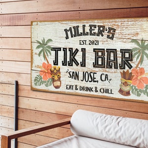 K&ouml;nnte beinhalten: Ein wei&szlig;es Holzschild mit dem Text "MILLER'S EST. 2021 TIKI BAR SAN JOSE, CA EAT & DRINK & CHILL" in schwarzer Schrift. Das Schild ist mit Palmen, Hibiskusbl&uuml;ten und Tiki-Masken verziert.