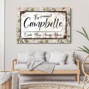 Op de afbeelding: Een rechthoekig familienaambord met de naam "The Campbells" in sierlijk schrift. Daaronder staan de namen "Linda, Steven, James, Nicole" vermeld. De achtergrond heeft een verweerde, vintage uitstraling met bloemaccenten.